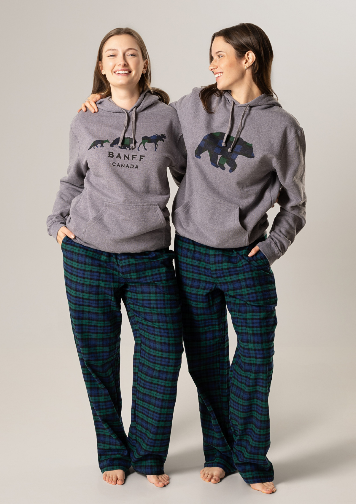 Two people wearing matching gray hoodies with a bear design and 'Banff Canada' text, paired with plaid pants on a plain background.