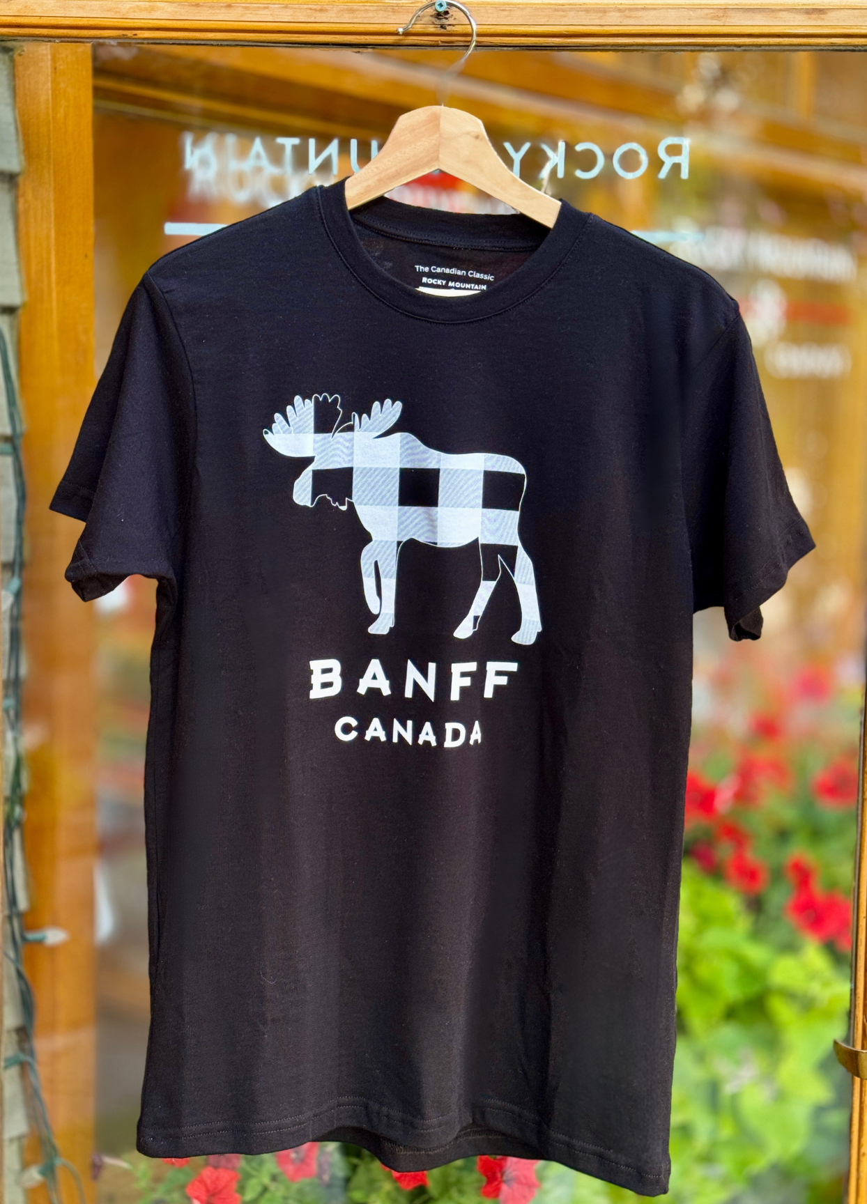 Black t-shirt with moose graphic and 'Banff Canada' text on a hanger in front of a store entrance.