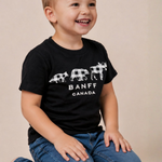A young boy smiling and looking to the side, wearing a black Banff Canada t-shirt with a white buffalo graphic and blue jeans, sitting against a plain beige background.