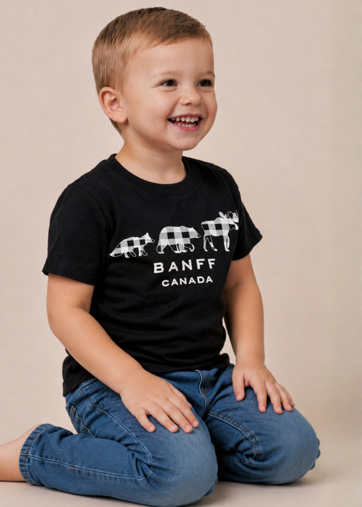 A young boy smiling and looking to the side, wearing a black Banff Canada t-shirt with a white buffalo graphic and blue jeans, sitting against a plain beige background.