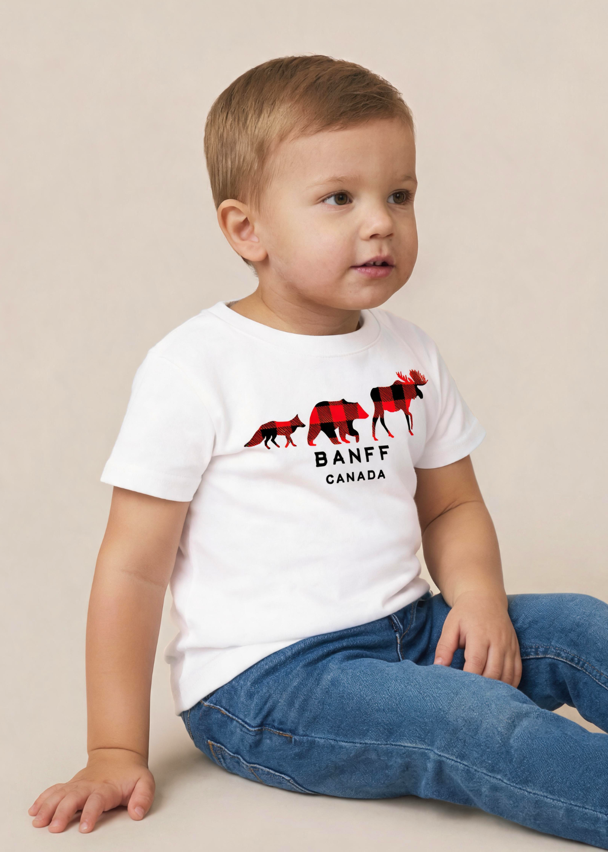 A toddler boy wearing a white short-sleeve t-shirt with a red and black moose graphic and the text 'BANFF CANADA', paired with blue jeans, sitting against a plain beige background.