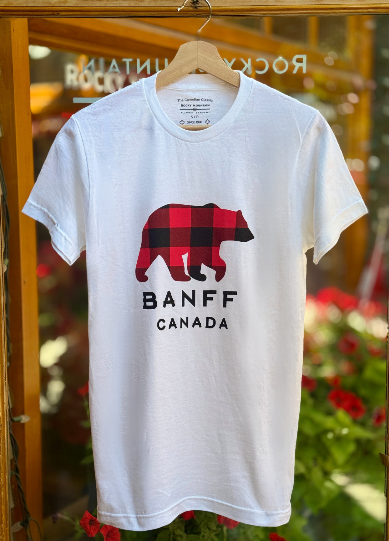 White t-shirt with a bear graphic and 'Banff Canada' text on a hanger against a blurred background.