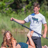 A woman sitting on a paddleboard wearing black watch shorts and a man standing next to her with a paddle, both smiling.