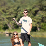 A woman sitting on a paddleboard wearing black watch shorts and a man standing next to her with a paddle, both smiling.