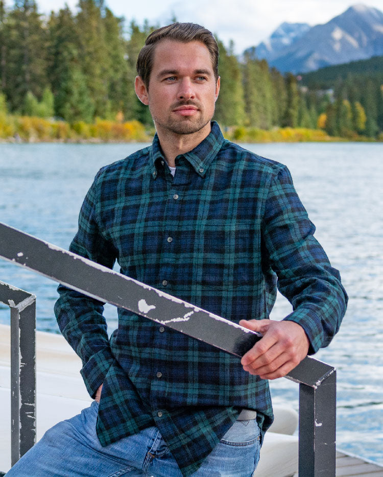 A man wearing a green plaid flannel shirt with a single breast pocket and two-button cuff, seated on a deck with a mountainous landscape in the background.