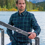 A man wearing a green plaid flannel shirt with a single breast pocket and two-button cuff, seated on a deck with a mountainous landscape in the background.