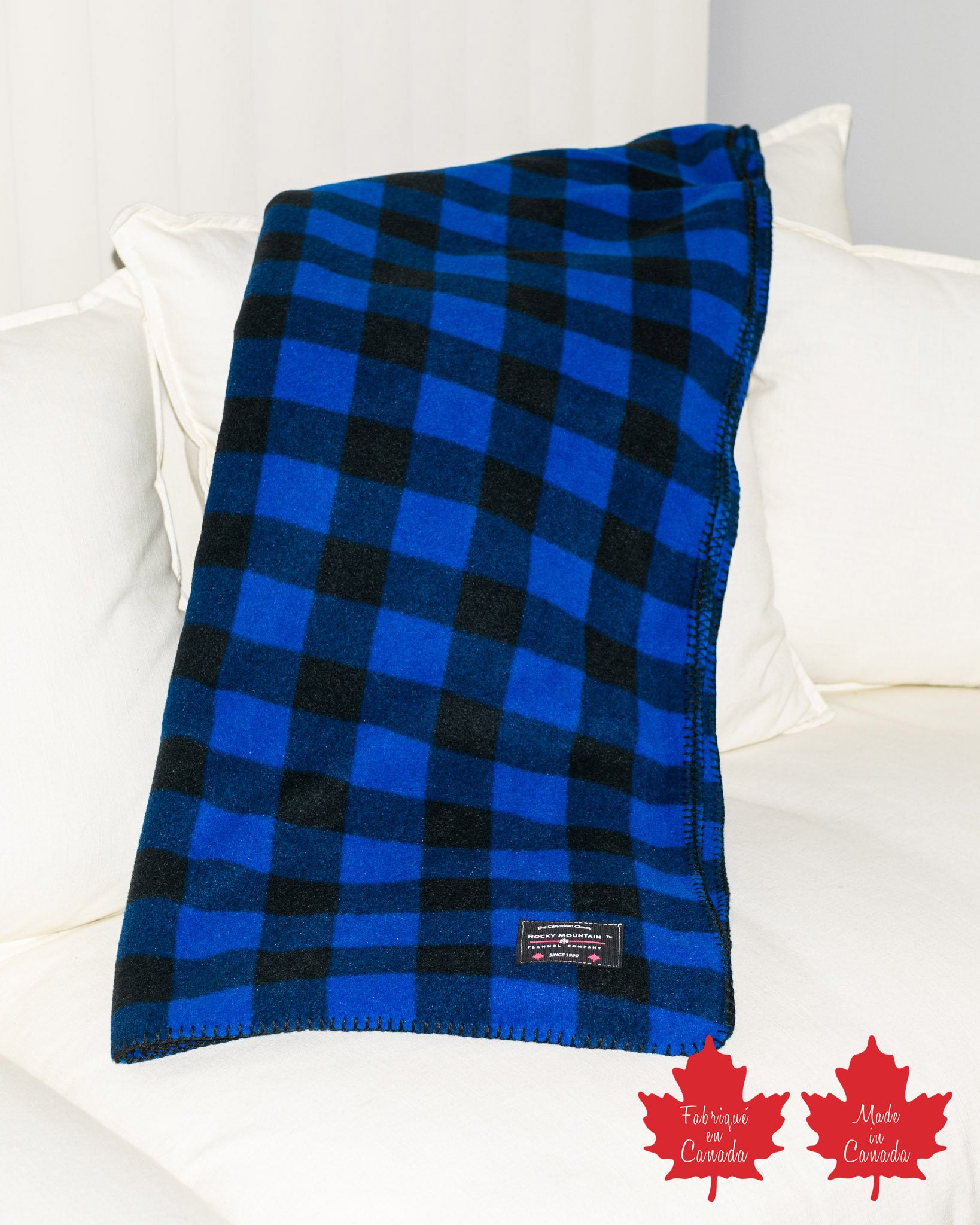 A blue and black checkered fleece blanket laid out on a white sofa, with a small branded tag visible.