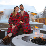 A man and a woman sitting by a fire, both wearing red and blue plaid kimono style flannel robes.